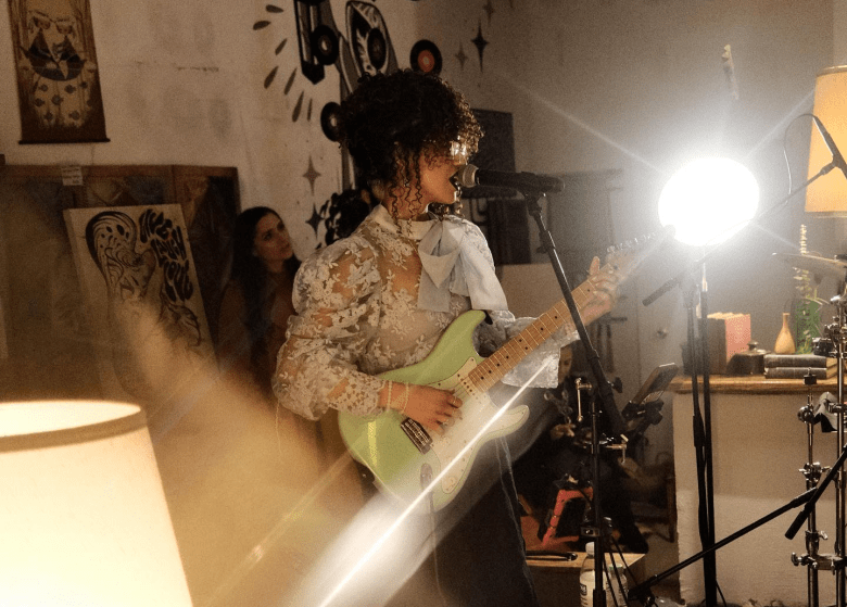A live performance photograph of songwriter SUNDE singing into a microphone while playing a seafoam green electric guitar. Captured in a medium profile shot from the right, SUNDE wears eyeglasses, dark curly hair tied up, and a high-collared light-colored top with sheer lace sleeves and a prominent neck bow. The indoor venue is brightly lit with warm stage lights and lens flares, with artwork on white walls and onlookers visible in the background.