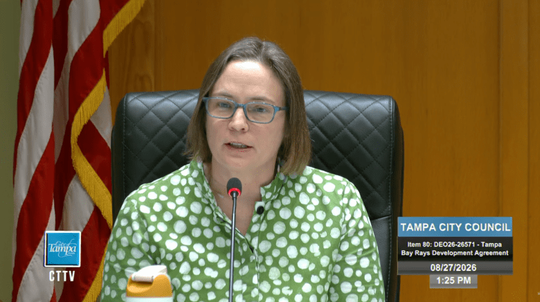 A screenshot from a YouTube broadcast showing Tampa City Councilwoman Lynn Hurtak speaking during a Tampa City Council meeting on August 27, 2026, at 1:25 PM. Captured in a medium shot, Councilwoman Hurtak sits in a black tufted leather chair speaking into a desktop microphone while addressing the council agenda regarding the Tampa Bay Rays Development Agreement. She wears rectangular eyeglasses and a green button-down shirt patterned with white polka dots, with the American flag visible behind her to the left and a live broadcast graphic overlay on the lower right.