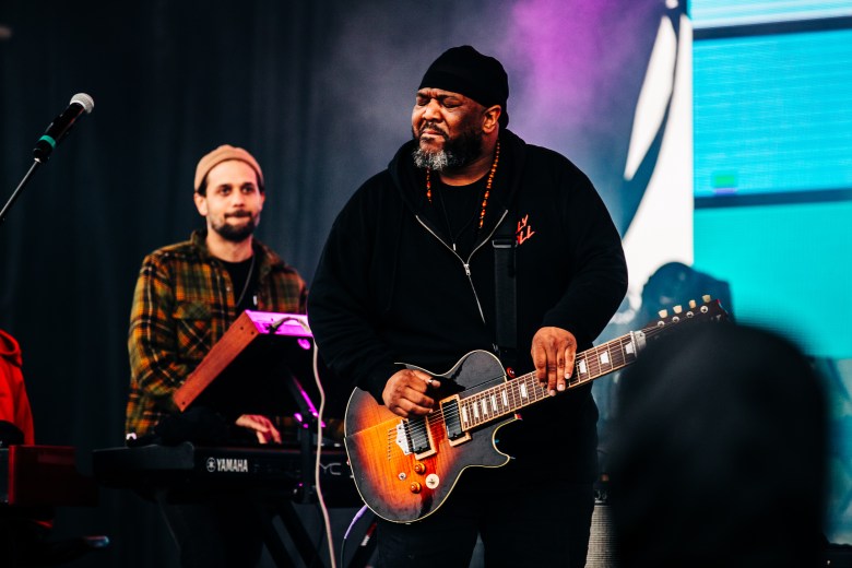 A concert photograph by Taylor Ysanne showing steel guitarist Roosevelt Collier performing live at the Gasparilla Music Festival in Julian B. Lane Riverfront Park in Tampa, Florida, on February 18, 2026. Captured in a medium shot under stage lighting, Collier stands center stage wearing a black beanie, black hoodie, and a dark beaded necklace while playing a sunburst single-cutaway electric guitar. Behind him to the left, a keyboardist wearing a tan beanie and plaid shirt plays a Yamaha synthesizer.