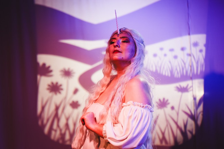 A theatrical performance photo by Dave Decker showing actor Jessica Jacobs performing in "The Last Unicorn" at The Fringe Theatre inside Kress Contemporary in Ybor City, Florida, on August 12, 2026. Captured in a medium close-up shot, Jacobs stands center stage under purple and warm stage lighting wearing a white off-the-shoulder peasant blouse, long light-colored hair, and a singular white unicorn horn attached to her forehead. She holds one hand near her chest while tilting her head back with closed eyes against a background illuminated by a shadow puppet projection of a floral field.
