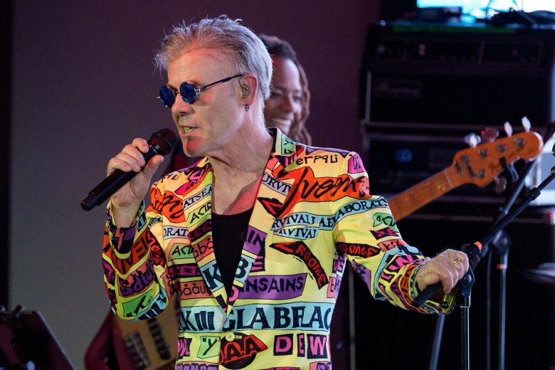 A photograph by Geoffrey Clowes showing musician Thomas Dolby performing live on stage in Las Vegas, Nevada, on July 25, 2026. Captured in a medium close-up shot under vibrant stage lighting, Dolby sings into a hand-held black microphone while wearing blue-tinted oval sunglasses and a colorful, pop-art style jacket covered in bold printed text graphics. In the background, a bass guitar with a sunburst finish and a smiling musician are visible on stage near performance equipment.