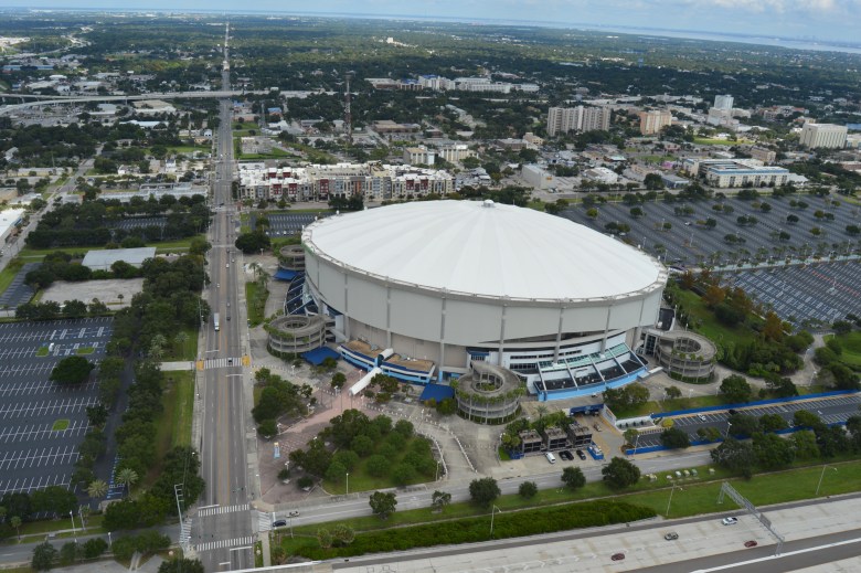 High-angle aerial photograph showing Tropicana Field in St. Petersburg, Florida, captured on October 2, 2015. The shot highlights the domed stadium's distinct tilted white roof structure surrounded by blue accent walkways and extensive parking lots, bordered by major roadways, surrounding commercial buildings, and green trees extending toward Tampa Bay in the background under a light blue sky.
