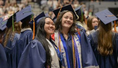 Two graduates in blue caps and gowns smile together at a commencement ceremony, wearing sashes and tassels.