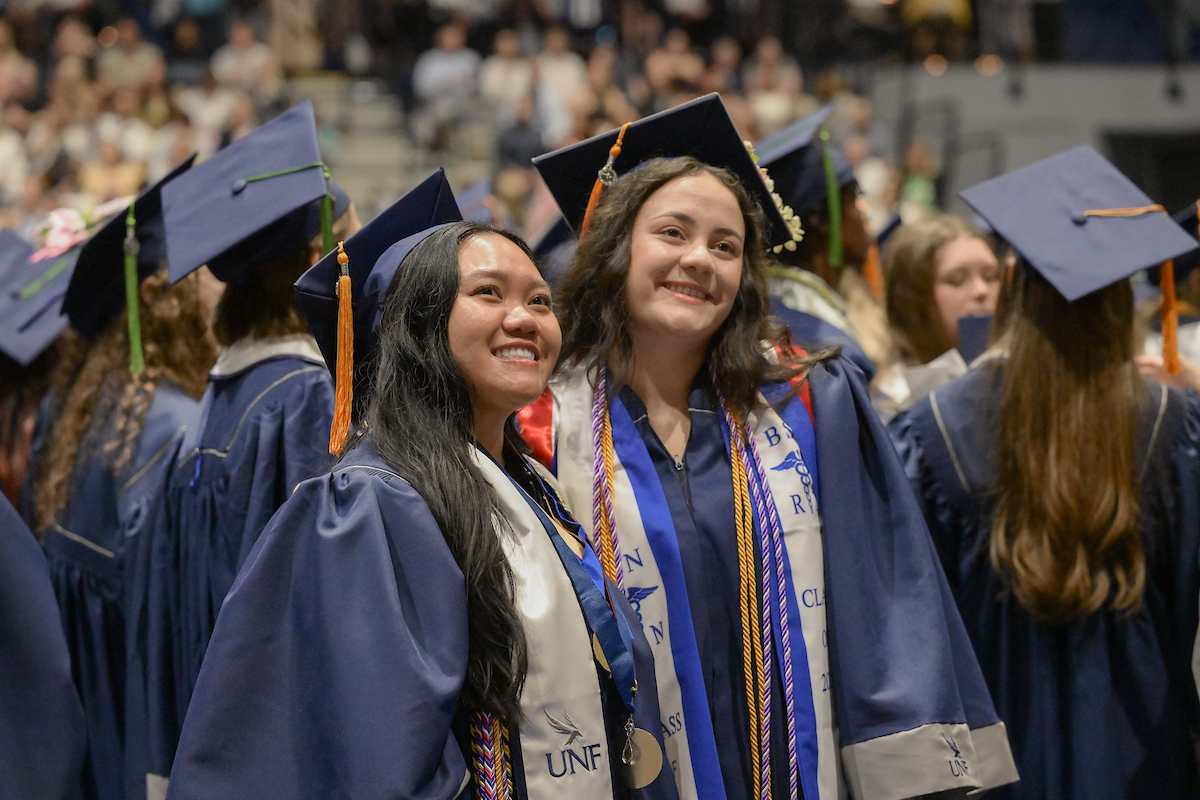 Two graduates in blue caps and gowns smile together at a commencement ceremony, wearing sashes and tassels.