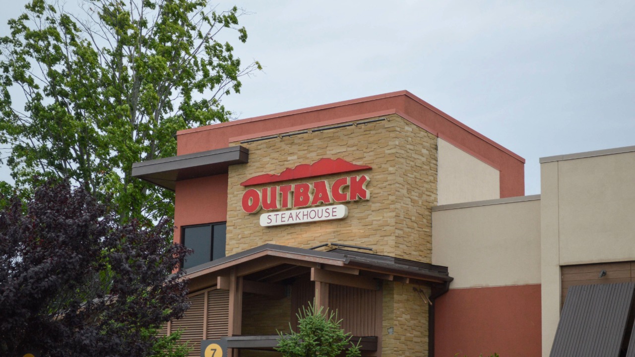 Exterior of an Outback Steakhouse restaurant with the chain’s red logo above the entrance.