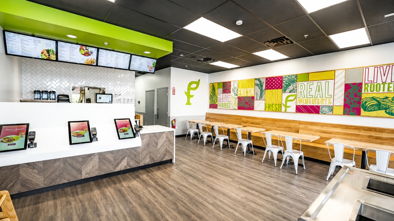 Interior of a SoFresh fast-casual restaurant with digital menu boards, ordering kiosks, green branding, seating area and wall mural.