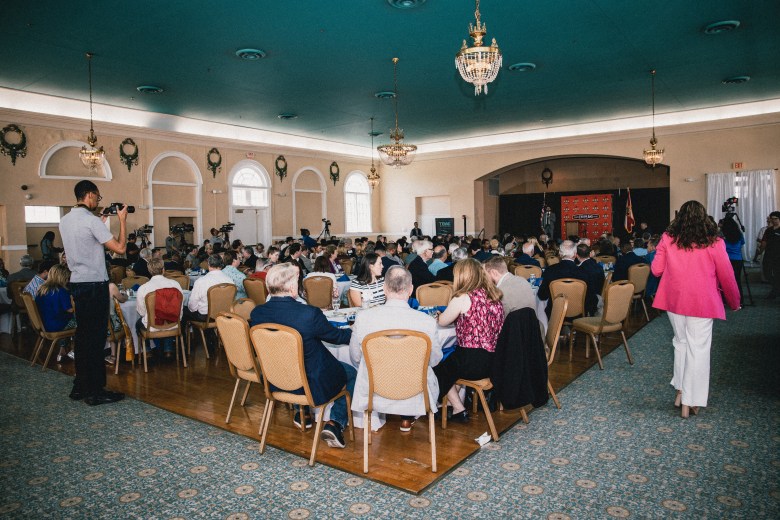 A wide-angle interior photograph by Dave Decker showing a crowded luncheon event hosted by the Tampa Tiger Bay Club at the historic Cuban Club in Ybor City, Florida, on April 17, 2026. Set inside a large ballroom with teal ceilings and chandelier lighting, numerous tables are filled with attendees seated for lunch while facing a stage adorned with orange backdrop banners at the far end of the room. A photographer stands on the left framing the crowd, while a speaker or guest in a bright pink jacket walks down the right aisle toward the stage.