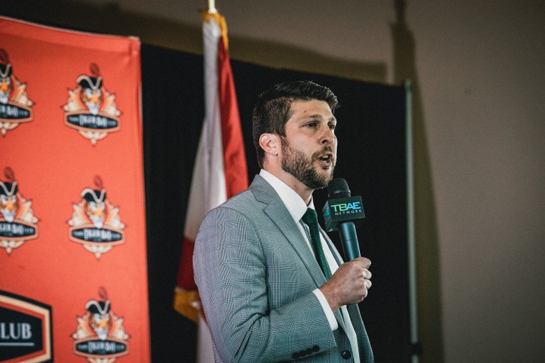 A photograph by Dave Decker showing James Uthmeier speaking at a Suncoast Tiger Bay Club event at the Cuban Club in Ybor City, Florida, on April 17, 2026. Positioned center-right in a medium profile shot, Uthmeier holds a microphone in his left hand while speaking, wearing a grey textured suit jacket, white dress shirt, and dark green tie. He features short dark hair and a groomed beard. In the background, a Florida state flag hangs behind him alongside an orange Tiger Bay Club step-and-repeat banner and a dark stage backdrop under indoor lighting.