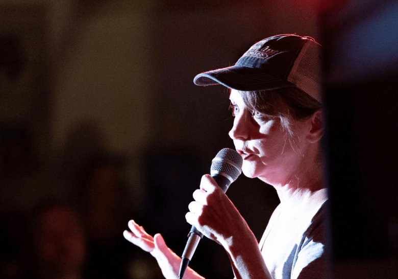 A close-up side-profile photograph of Virginia Pelley speaking into a handheld microphone during a live stage event. Illuminated by a strong white spotlight on her face with subtle red stage ambient lighting, Pelley holds the silver condenser microphone in her right hand while gesturing outwards with her open left hand. She wears a dark baseball cap with a mesh back and curved brim, along with a casual t-shirt, set against a dark, out-of-focus background.