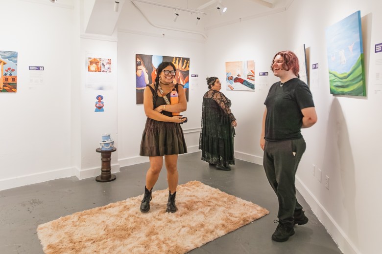 A photograph showing attendees viewing art inside a brightly lit gallery during the Ybor Arts Tour in Ybor City, Florida, on February 19, 2026. Captured in a wide shot against white gallery walls and polished grey concrete floors, two visitors chat near the center on a beige faux-fur area rug: a person on the left wearing black-framed glasses, a dark sleeveless dress, and black boots smiles, while a person on the right in a black t-shirt and dark trousers talks beside framed paintings, including a landscape artwork on the far right wall. A third guest in a long black lace dress views artwork further back in the gallery under track lighting