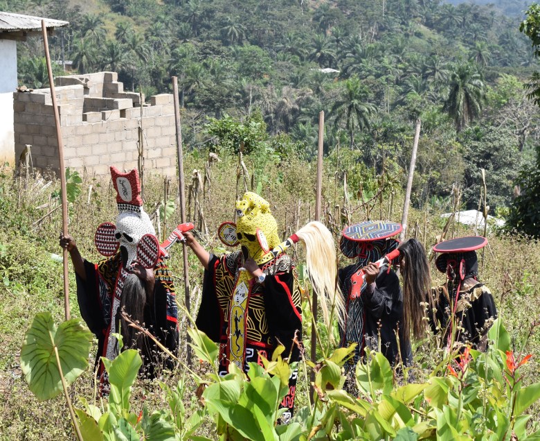 An outdoor photograph showing Hervé Youmbi's Tso Scream Mask and Tso Scream Leopard Mask performing at the Nka’a Kossié society succession ceremony at the Fondati Chieftaincy on December 3, 2022. Captured in a medium-wide shot on a grassy hillside, four masked masqueraders walk single-file from left to right holding long wooden staves and light-colored horsehair flywhisks. On the left, the Tso Scream Mask wearer sports the white beaded Scream-style face mask and a red-and-white crown structure, followed by the yellow-masked Tso Scream Leopard Mask wearer and two additional masqueraders in dark headpieces and embroidered tunics, set against a background of green hills and a concrete building under natural daylight.
