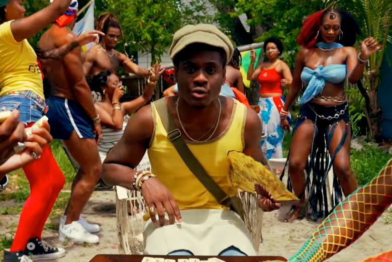A screenshot from the music video for Beanie's single "Beanie Baby." The main subject, musician Beanie, sits centered in a woven chair wearing a yellow tank top, light cap, silver chain necklaces, an olive crossbody strap, and beaded bracelets, holding a yellow patterned fan while looking into the camera. Around them, dancers perform outdoors under sunny lighting, including a person on the left in a yellow shirt and red tights, a shirtless dancer in blue athletic shorts, and a dancer on the right in a light blue top with split black fringe shorts against a lush green tree backdrop.