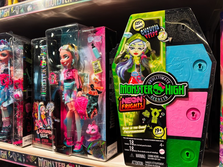 A photograph showing Monster High dolls displayed on a store shelf in Lviv, Ukraine, on February 16, 2025. Positioned center-right, a black coffin-shaped box from the "MONSTER HIGH NEON FRIGHTS" series features neon green, pink, and blue secret compartment doors with keyhole slots, alongside Ukrainian-language promotional stickers. To its left, boxed Monster High fashion dolls with colorful hair and outfits sit packaged in clear plastic window boxes, lined up under retail store lighting.