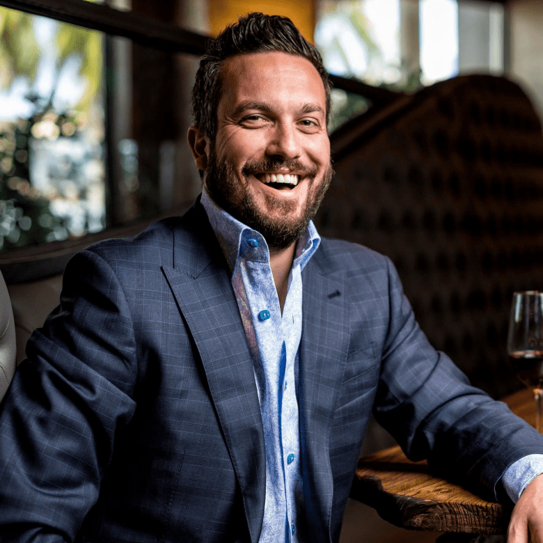 A portrait photograph of celebrity chef and restaurateur Fabio Viviani sitting at a wooden restaurant table. Positioned center frame in a medium close-up shot, Viviani smiles broadly toward the camera, featuring dark styled hair and a full groomed beard. He wears a dark grey plaid suit jacket over an unbuttoned light blue patterned dress shirt with turquoise buttons. Behind him, warm interior lighting illuminates a stylish restaurant dining room with dark leather booth seating and window reflections in the soft-focus background.
