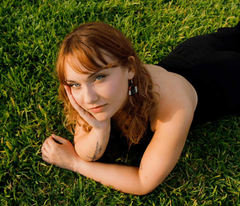A photograph by Emma Hintz showing Tampa Bay area DJ and artist Delaney Staack lying down on a green grassy lawn. Captured in a top-down, high-angle portrait, Staack rests her chin in her right hand, revealing a small tattoo on her forearm, and looks toward the camera. She features auburn hair with bangs, light eyes, dangling statement earrings, and wears a black strapless top, lit by warm, angled sunlight.