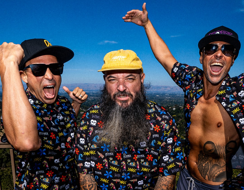 A promotional group photograph showing three members of the Hawaiian reggae-rock band Pepper posing enthusiastically outdoors under a bright blue sky. On the left, vocalist and bassist Bret Bollinger wears dark sunglasses, a black baseball cap, and a short-sleeved button-down shirt patterned with skulls and flowers while making a triumphant fist. In the center, drummer Yesod Williams wears a yellow baseball cap, a long greying beard, and a matching patterned short-sleeved shirt with his arms raised. On the right, guitarist and vocalist Kaleo Wassman wears sunglasses, a black cap, and an unbuttoned matching floral skull shirt exposing his torso and abdominal tattoo while raising his hands with a wide open-mouthed expression.