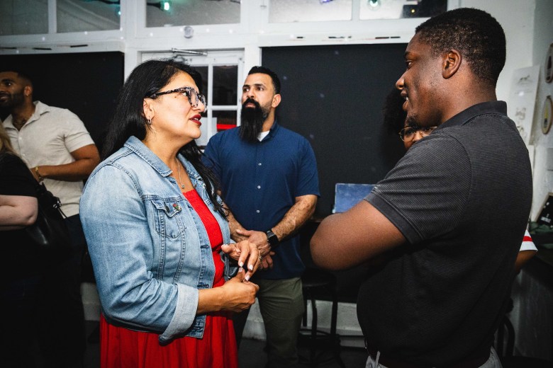 A photograph by Dave Decker showing U.S. Rep. Rashida Tlaib on the left and Elijah Manley on the right conversing at Equality Park in Broward County, Florida, on August 14, 2026. Captured in a medium shot inside an event venue, Tlaib wears black-framed glasses, a denim jacket over a bright red dress, and stands facing right while speaking to Manley, who stands in profile facing left wearing a dark short-sleeved polo shirt with his arms crossed. In the background between them, a person with a dark beard and blue button-down shirt looks on under warm indoor lighting.