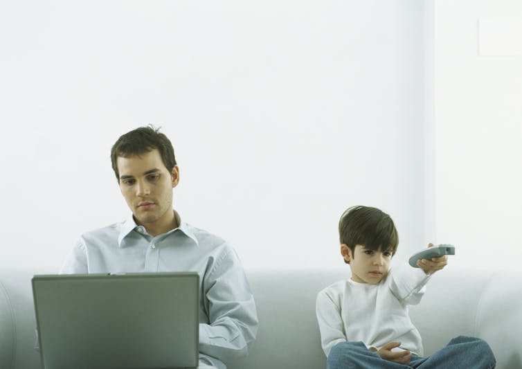 A man sits in front of a computer screen while a child holding a remote control sits next to him.