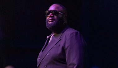 Rick Ross in a dark suit and sunglasses stands on stage with a confident expression, under purple lighting.