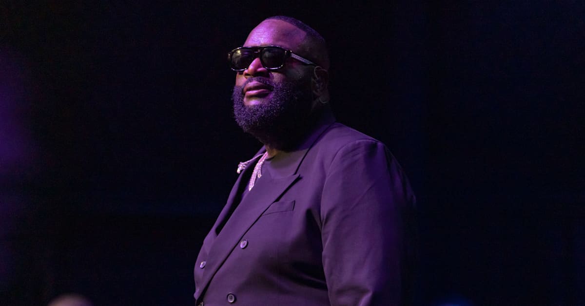 Rick Ross in a dark suit and sunglasses stands on stage with a confident expression, under purple lighting.