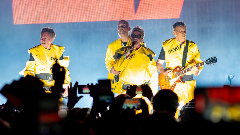 A concert photograph showing band members of Devo—(L-R) Gerald Casale, Josh Hager, Mark Mothersbaugh, and Bob Mothersbaugh—performing live at the Eventim Apollo in London, England, on August 19, 2023. Captured in a wide shot from within the crowd, the four musicians stand on stage wearing their signature bright yellow radiation-style protective suits with "DEVO" printed across the chest, dark glasses, and black shoulder straps. Mark Mothersbaugh sings into a microphone at center stage while Bob Mothersbaugh plays a sunburst electric guitar on the right, as audience members hold up smartphone screens in silhouette in the foreground under bright stage backlighting.
