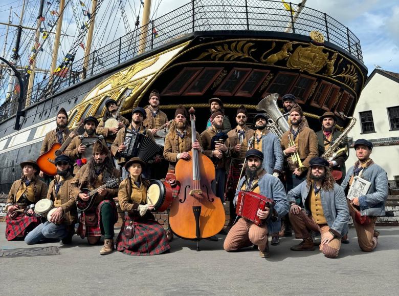 A promotional group photograph of the musical ensemble Old Time Sailors posing outdoors in front of a historic, ornate wooden sailing ship. Captured in a wide shot, over a dozen band members stand and kneel together in traditional 19th-century maritime attire, including newsboy caps, brown jackets, waistcoats, and trousers. The musicians hold and display various acoustic instruments, including double bass, accordions, guitars, violins, a tuba, a trumpet, washboards, and hand drums. The stern of the black and gold decorated ship towers behind them under a bright, partly cloudy sky in natural daylight.