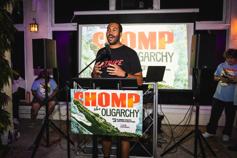 A photograph of St. Petersburg City Councilman Richie Floyd speaking at an event at Equality Park in Broward County, Florida, on August 14, 2026. Captured in a full-length shot inside a dimly lit venue under soft accent lighting, Floyd stands at a black podium holding a microphone while addressing the room. The podium displays a sign reading "CHOMP the OLIGARCHY" above an illustration of an alligator. Floyd wears a dark t-shirt featuring red text above his shorts, set against a large projection screen in the background displaying the same "CHOMP THE OLIGARCHY" graphic, with attendees seated on either side of the stage.