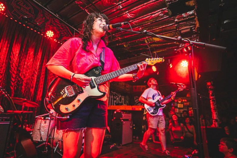 A concert photograph showing Elizabeth Stokes of the New Zealand indie rock band The Beths performing live on stage at Crowbar in Ybor City, Florida, on June 21, 2023. Positioned center-right in a medium three-quarter shot, Stokes sings into a stand-mounted microphone while playing a dark electric guitar with a white pickguard. She wears a bright pink short-sleeved collared shirt, dark denim shorts, and has dark shoulder-length hair with bangs. The stage is bathed in deep red performance lighting, with red curtains, stage lights, a drum kit, and a Pabst Blue Ribbon banner visible in the background.