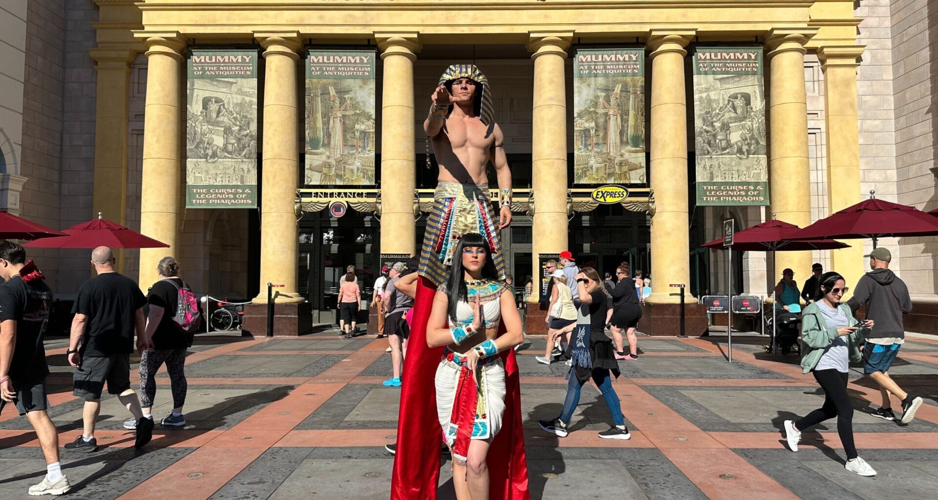 Two people in Egyptian costumes pose by a museum, like actors from Revenge of the Mummy Ride, as crowds stroll in sunlight.