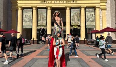 Two people in Egyptian costumes pose by a museum, like actors from Revenge of the Mummy Ride, as crowds stroll in sunlight.