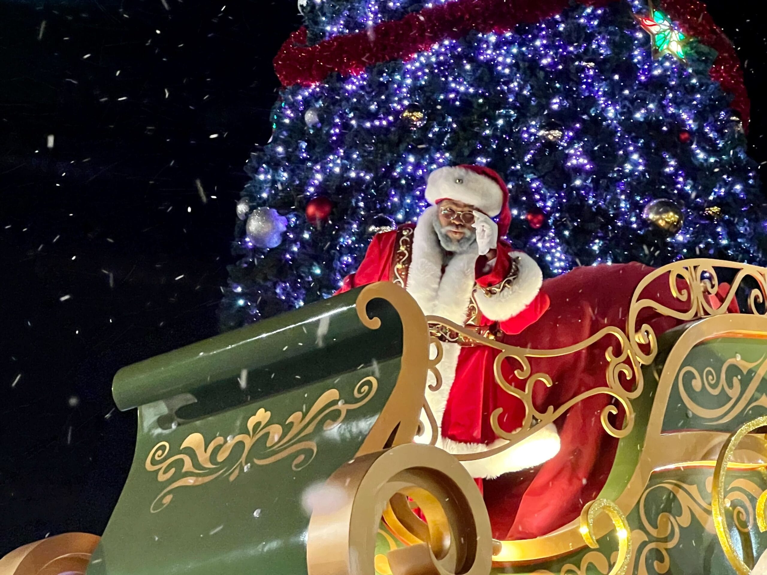 Santa Claus stands in a decorated sleigh in front of a large, lit Christmas tree, with falling snow around.