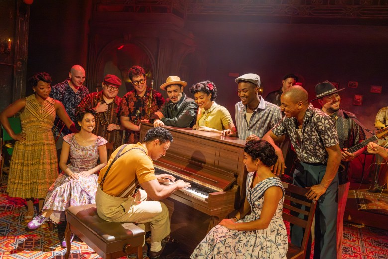 A stage production photograph by Matthew Murphy showing The North American Tour company of Buena Vista Social Club. Captured in a wide shot under warm, dramatic stage lighting, a musician seated on a bench plays an upright piano while surrounded by ensemble cast members. To his right, a seated performer in a pattern dress looks on intently, while other cast members dressed in mid-century attire—including patterned shirts, fedoras, flat caps, and vintage dresses—stand and gather around the piano leaning in, singing, and watching the performance against an ornate, carpeted stage set.
