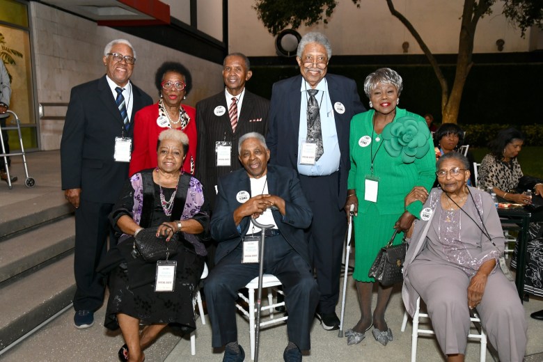A group portrait of civil rights pioneers and members of the 1960 Tampa Woolworth sit-ins outside the Franklin Exchange and Wilson buildings in downtown Tampa on Nov. 17, 2025. Seven well-dressed elderly individuals pose outdoors in front of a modern building entrance. Four men and one woman stand in the back row wearing formal suits, dark blazers, a red jacket, and a bright green dress suit with a large fabric flower accent. Seated in front on white folding chairs are three individuals, including a man in a dark suit holding a walking cane in the center and a woman on the right in a grey outfit. Most wear circular white "I SAT / I STAND" pin-back buttons and event credential lanyards.