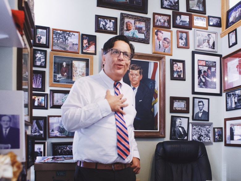 An indoor medium shot photograph by Michael Johnson showing Tampa City Councilman Luis Viera at Tampa City Hall in Tampa, Florida on June 23, 2026. Standing centrally in his office, Viera wears dark-framed glasses, a white long-sleeved dress shirt, a brown belt, and a blue, red, and white diagonally striped tie while holding his right hand over his heart and looking slightly off-camera to the right. The office wall behind him is completely covered with framed historical portraits and political photographs, including prominent framed portraits of John F. Kennedy and Abraham Lincoln, alongside a black leather office chair in the background.