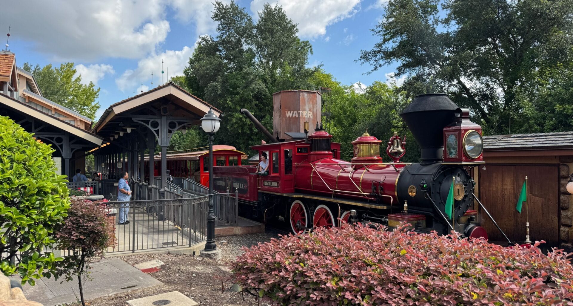 A bright red vintage steam train, like the Walt Disney World Railroad, waits at a station amid trees and greenery.