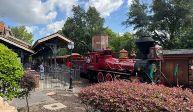 A bright red vintage steam train, like the Walt Disney World Railroad, waits at a station amid trees and greenery.
