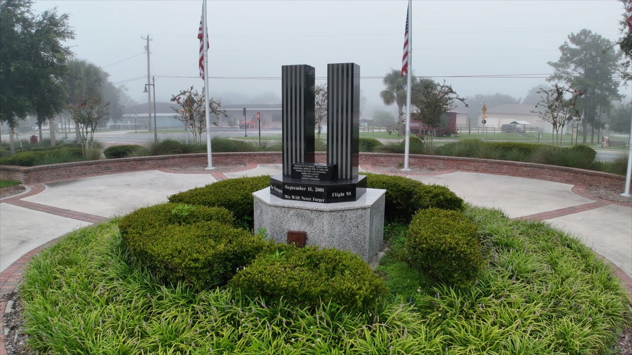 The 9/11 Memorial in Inverness is located in the Depot Pavilion area at Liberty Park. Two dark columns stand in the center on a large pedestal, with American flags marking the outer circle.