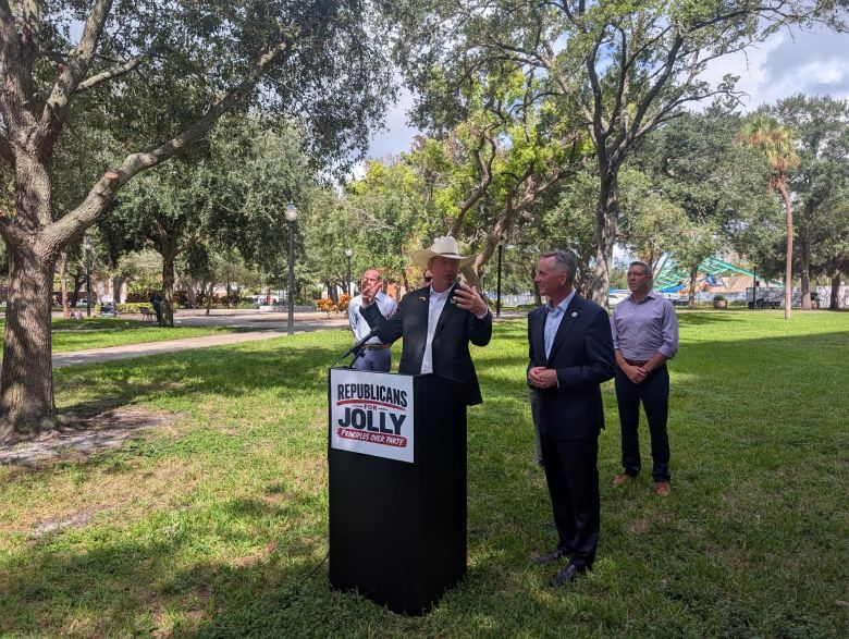 Florida Agriculture Commissioner Wilton Simpson wearing a cowboy hat speaks at an outdoor press conference in a tree-shaded park alongside government officials.