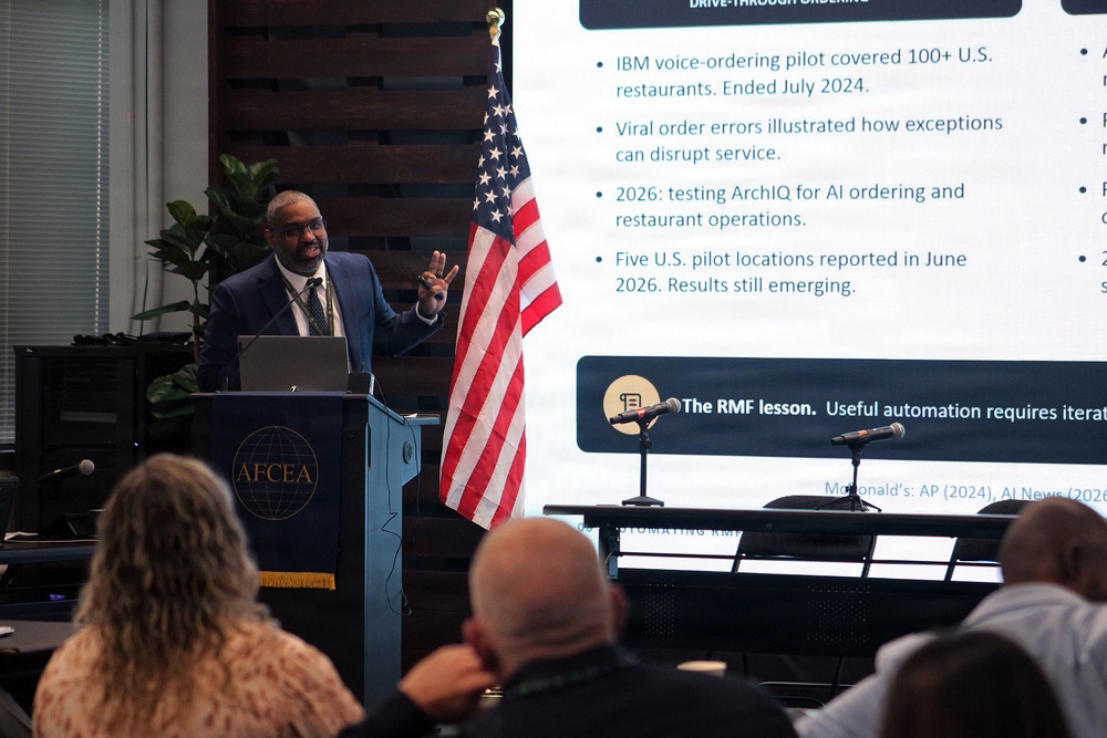 A man in a blue suit is presenting at an AFCEA podium before an audience, with a large projection screen behind him displaying content about AI drive-through ordering systems, including references to IBM voice-ordering pilots and McDonald