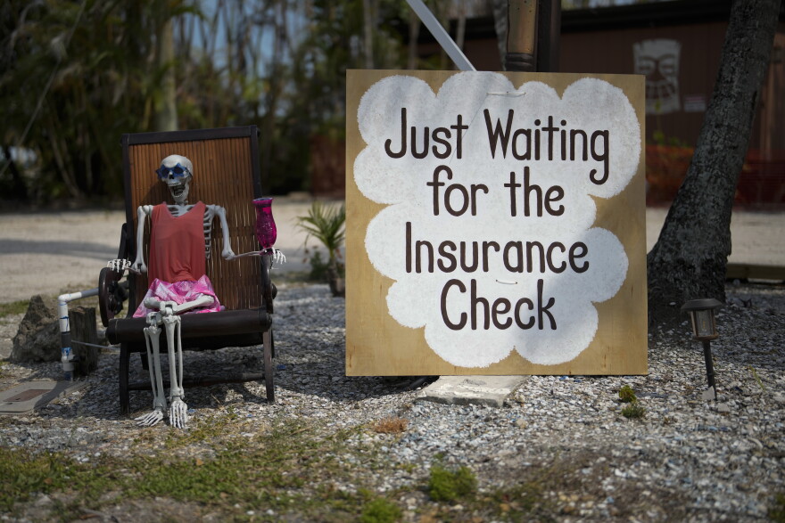 A skeleton in sunglasses sits beside a sign reading: "Just waiting for the insurance check," outside the closed Kona Kai Motel on Sanibel Island, Fla., Thursday,