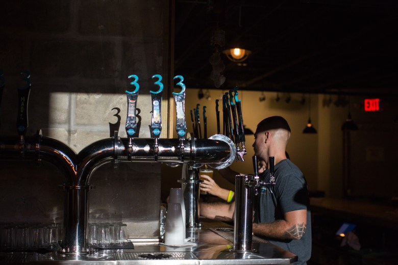 A photograph of a bartender pouring a beer at 3 Daughters Brewing St. Petersburg Florida. Captured in profile view inside a dimly lit taproom, the bartender wearing a grey t-shirt, backwards black baseball cap, and forearm tattoo reaches toward a stainless steel draft beer tower fitted with custom tap handles featuring blue number "3" logos. In the background, hanging pendant lights, a red "EXIT" sign, and concrete block walls frame the bar area.