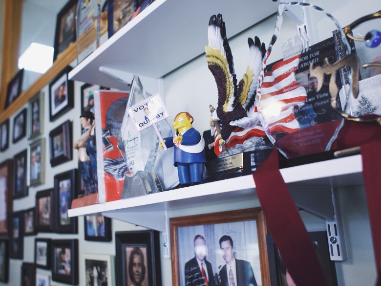 An indoor close-up photograph by Michael Johnson showing memorabilia on display on white shelving inside a City Hall office, featuring a figurine of Mayor 'Diamond Joe' Quimby at Tampa City Hall in Tampa, Florida on June 23, 2026. Positioned in the center of the shelf, the blue-suited figurine of The Simpsons character holds a small white sign reading "VOTE FOR QUIMBY." Beside it stand an American flag plaque, a bald eagle statue, and framed photographs, while the background wall on the left is lined with dense, framed portraits under bright indoor lighting.