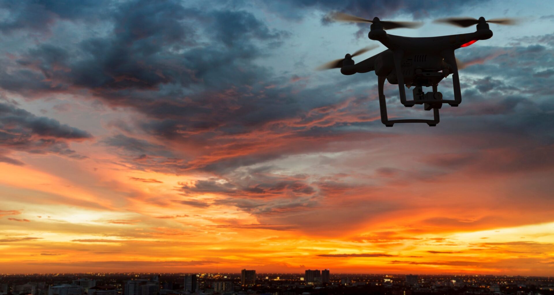 Drone flying above a city skyline at sunset beneath a cloudy sky.
