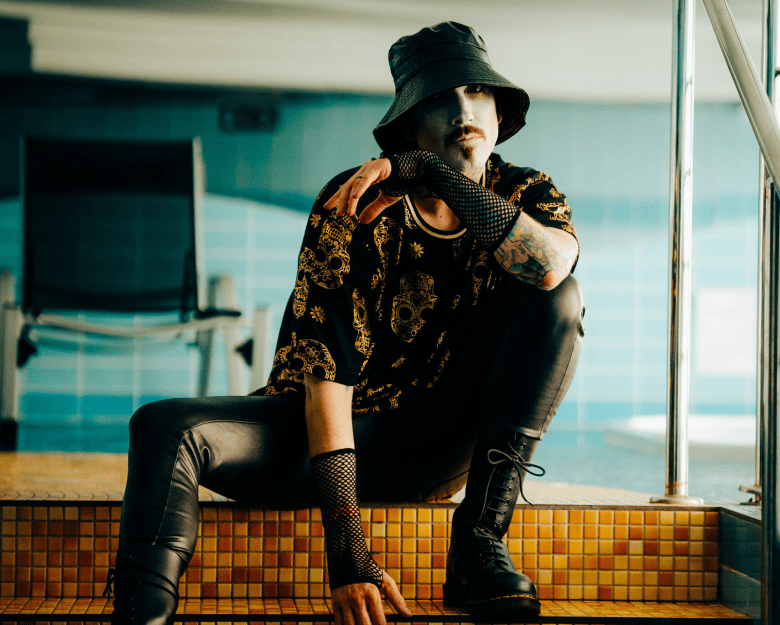 A promotional portrait photograph showing electronic musician Daniel Graves of Aesthetic Perfection. Perched on tiled steps near an indoor swimming pool, Graves wears a black bucket hat, a dark t-shirt with gold skull-and-floral patterns over a black undershirt, black mesh fingerless arm sleeves, fitted black leather pants, and black lace-up combat boots. He crouches with his right hand resting on the steps and his left elbow rested on his knee under soft indoor lighting.