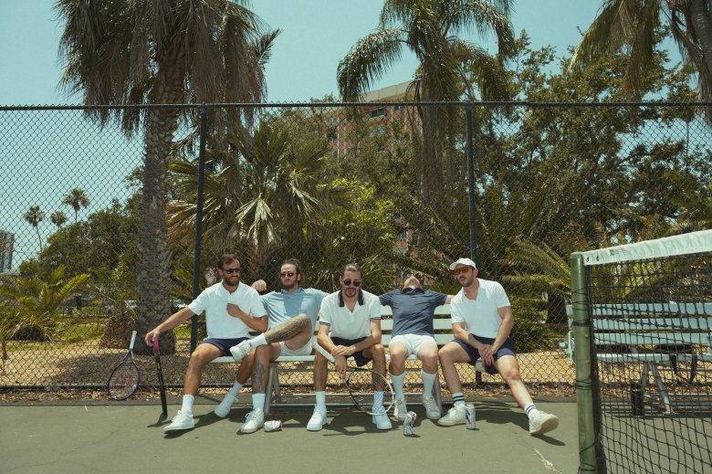 A promotional group portrait of funk-rock band Ajeva sitting together on a court bench at an outdoor tennis court. The five band members are seated side-by-side in athletic tennis wear—including white collared shirts, dark shorts, white crew socks, white sneakers, and a white baseball cap on the far right. Behind them, a chain-link fence separates the court from lush tropical palm trees and greenery under bright, direct sunlight, with a tennis net visible in the right foreground frame.