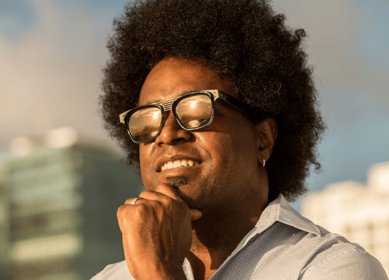A close-up outdoor portrait of artist Alexandre Arrechea. Smiling in warm sunlight, Arrechea wears gold reflective sunglasses, a white short-sleeved collared shirt, and a small silver hoop earring, with his right hand resting under his chin. He has a full afro hairstyle and a neatly trimmed goatee, set against a soft-focus background of city buildings and a bright blue sky.