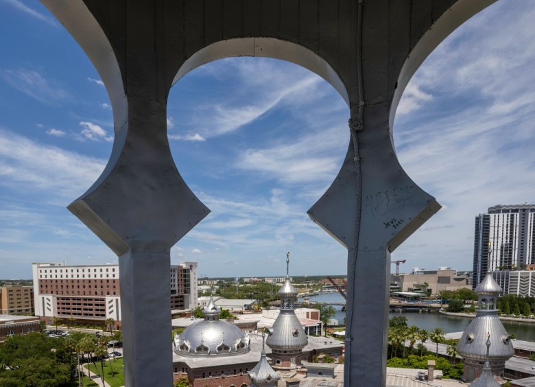 A wide elevated architectural photograph by Amy Martz showing Downtown Tampa and the University of Tampa as seen from inside one of the school's iconic minarets. Framed from behind white keyhole archway pillars inside the elevated minaret, light graffiti inscriptions reading "Audrey Till '25" and "Jevs 2023" are visible etched into the right column. In the foreground below, silver stainless-steel domes and spires top the brick campus roofline, while the Hillsborough River, downtown high-rise buildings, and surrounding city streets stretch out into the background under a bright blue sky filled with wispy white clouds.