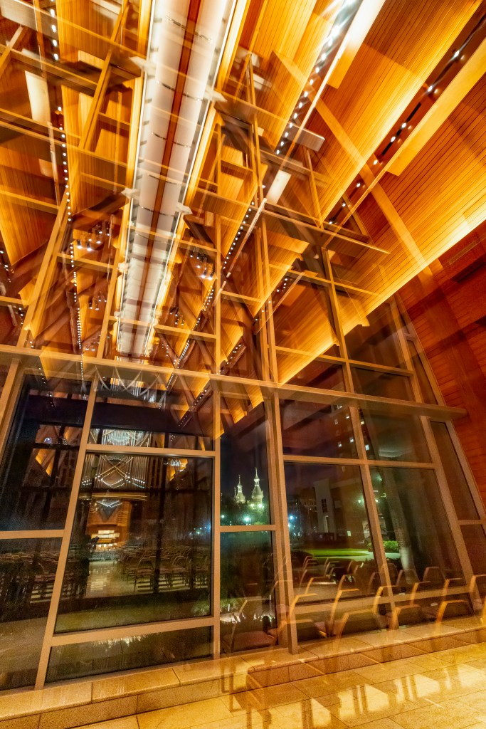 A vertical interior architectural photograph by Amy Martz showing Inside the Sykes Chapel and Center for Faith and Values at UTampa in Tampa, Florida. Looking up toward soaring warm wooden vaulted ceiling beams illuminated by linear track lights, the shot captures a large glass wall facade reflecting the interior pipe organ and wooden pews. Through the floor-to-ceiling glass windows at night, the lit minarets and illuminated towers of Plant Hall are visible across campus in the distance.