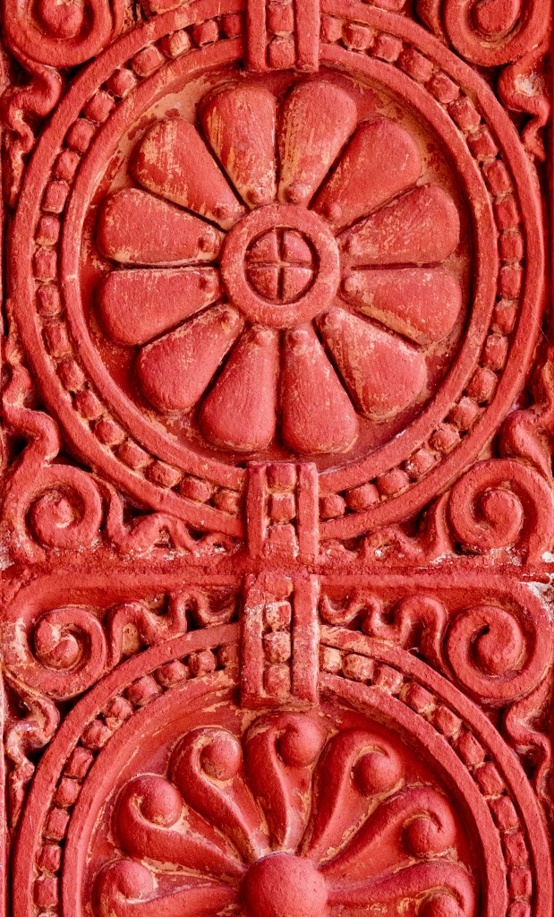 An architectural detail photograph by Amy Martz showing Flowers like this are all over the University of Tampa. Captured in a tight vertical macro shot, an intricate terracotta flower relief carving in deep red-orange clay forms a symmetrical floral rosette surrounded by beaded circular borders, ornate scrollwork motifs, and raised geometric patterns on the exterior facade of Plant Hall.
