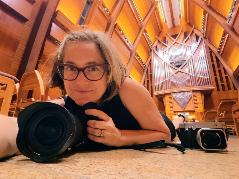 A low-angle self-portrait photograph showing photographer Amy Martz inside the Sykes Chapel and Center for Faith and Values at UTampa in Tampa, Florida. Lying prone on the chapel's polished stone floor with her professional Canon camera lens pointed toward the foreground, Martz wears dark-rimmed glasses and a dark sleeveless dress while resting her chin in her hand and smiling slightly at the camera. A silver Leica rangefinder camera rests on the floor beside her, positioned before the soaring warm-wood vaulted ceilings, exposed timber arches, and tall pipe organ of the chapel interior under warm ambient light.